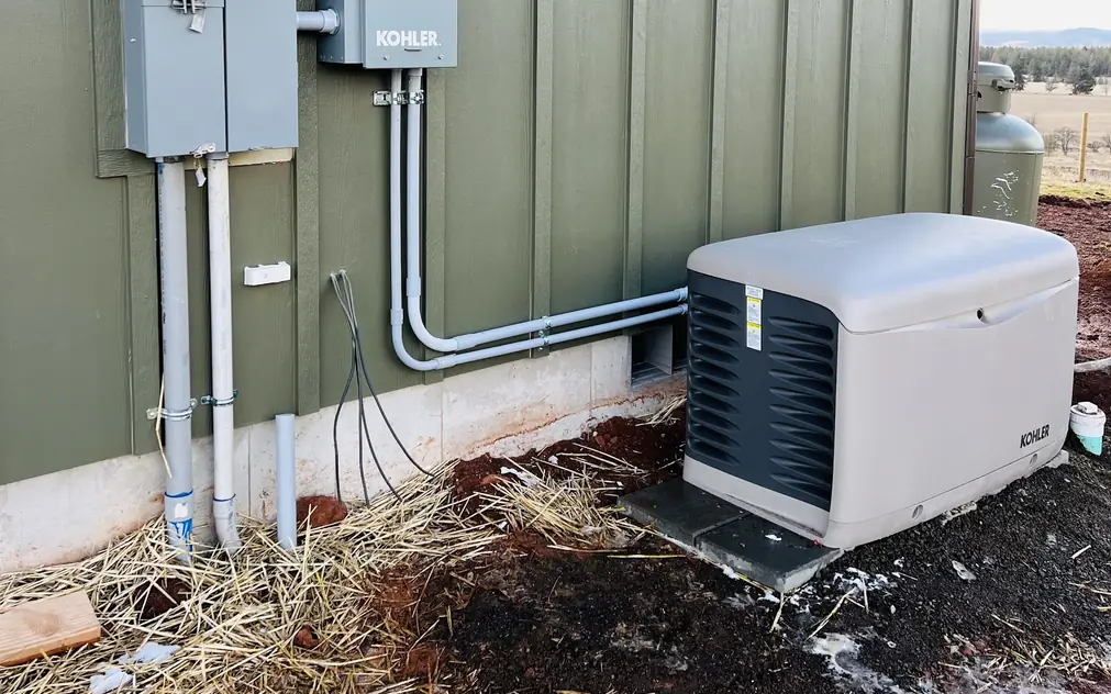 A standby generator and transfer switch installed beside a green board-and-batten house